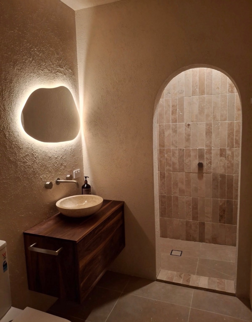 Warm textured bathroom with an illuminated organic mirror and an arched tumbled-stone shower
